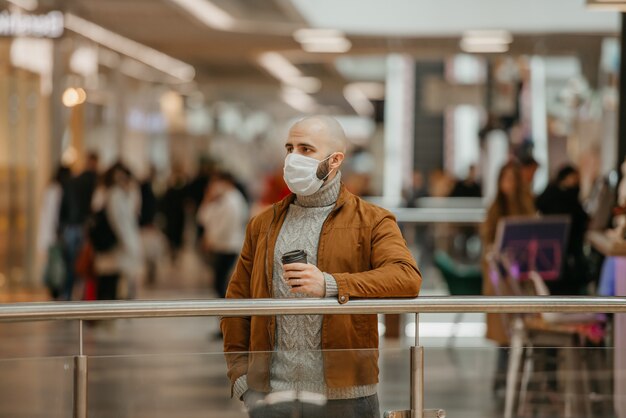 Un uomo con una maschera medica per evitare la diffusione del coronavirus tiene in mano una tazza di caffè mentre aspetta nel centro commerciale. Un ragazzo calvo con una mascherina chirurgica sta mantenendo le distanze sociali.