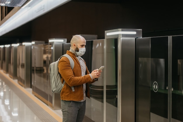 Un uomo con una maschera medica sta leggendo le notizie su uno smartphone e sta fissando di lato mentre aspetta un treno alla piattaforma della metropolitana. Un ragazzo calvo con una mascherina chirurgica sta mantenendo le distanze sociali.