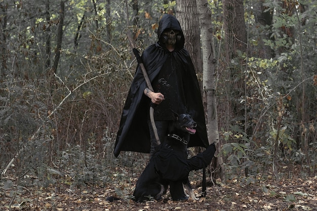 Un uomo con una maschera spaventosa e vestiti neri con un cane nero in una cupa foresta.un'immagine per Halloween e il giorno dei morti.