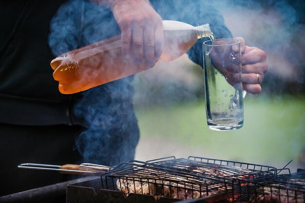 un uomo cucina la carne alla griglia e versa birra schiumosa fredda da una bottiglia in un bicchiere in natura