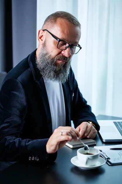 Un uomo d'affari adulto sta lavorando a un nuovo progetto e sta guardando i grafici di crescita delle azioni. Si siede al tavolo vicino alla grande finestra. Guarda lo schermo del laptop e beve caffè.