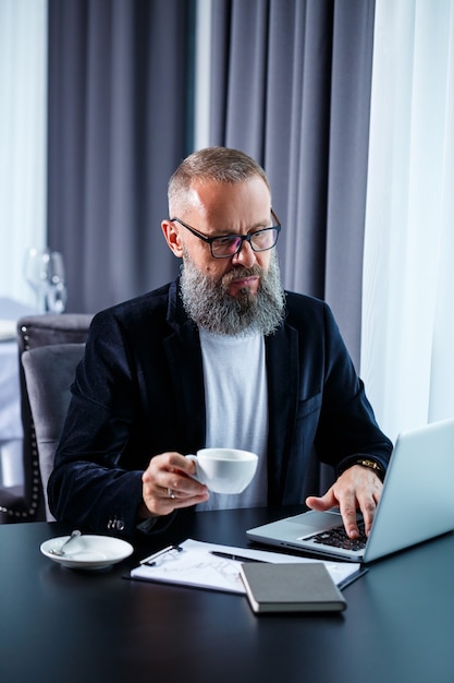 Un uomo d'affari adulto sta lavorando a un nuovo progetto e sta guardando i grafici di crescita delle azioni. Si siede al tavolo vicino alla grande finestra. Guarda lo schermo del laptop e beve caffè.