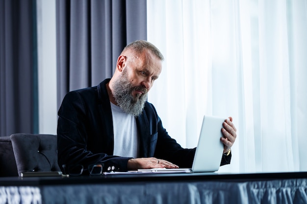 Un uomo d'affari adulto sta lavorando a un nuovo progetto e sta guardando i grafici di crescita delle azioni. Si siede al tavolo vicino alla grande finestra. Guarda lo schermo del laptop e sorride