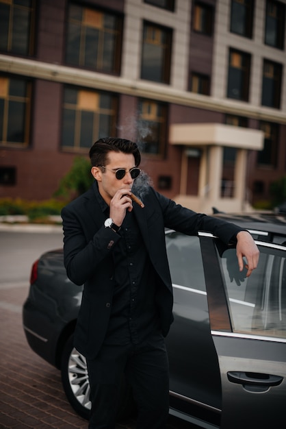 Un uomo d'affari alla moda fuma sigari vicino a un'auto di lusso. Moda e affari.