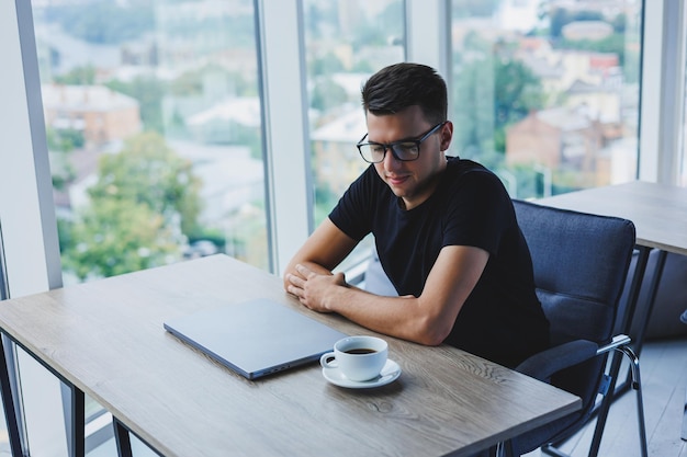 Un uomo d'affari allegro con gli occhiali si siede alla sua scrivania lavorando con un computer portatile in ufficio Un uomo d'affari si siede e lavora su un computer portatile e beve caffè Iniziare la giornata in ufficio