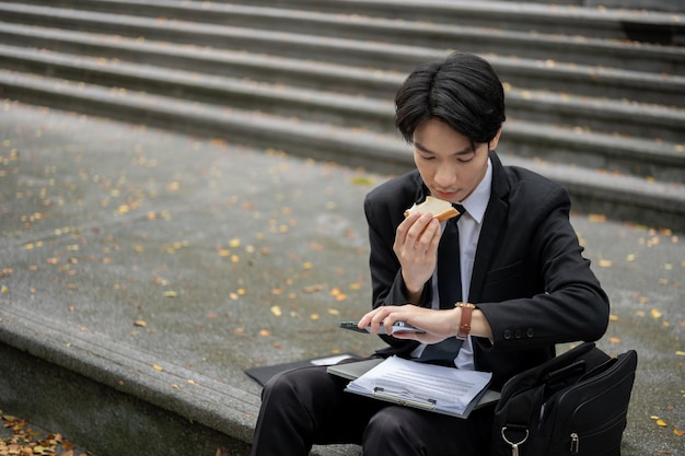 Un uomo d'affari asiatico impegnato sta controllando l'ora sul suo orologio e sta mangiando un panino su una scala