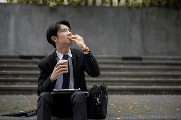 Un uomo d'affari asiatico occupato, stanco e affamato che mangia un panino su una scala