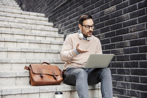 Un uomo d'affari casual intelligente si siede sulle scale all'aperto e ha una chiamata in conferenza