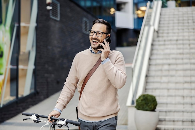 Un uomo d'affari casual sorridente sta spingendo la sua bicicletta per strada e sta parlando al telefono