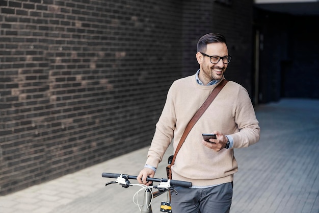 Un uomo d'affari casuale felice che spinge una bici sulla via e che utilizza un telefono