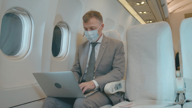 Un uomo d'affari caucasico sta lavorando con il computer portatile in aereo, indossando una maschera protettiva per la protezione covid-19 durante il viaggio