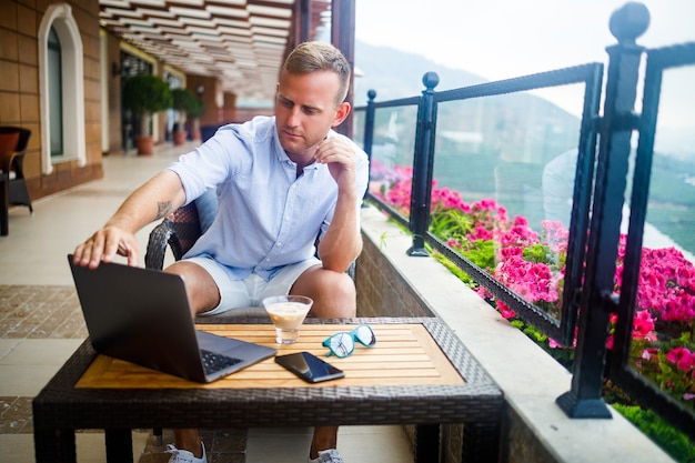 Un uomo d'affari di successo è seduto a un tavolo con un laptop, sta lavorando in vacanza. Vacanze e lavoro a distanza. Il ragazzo è seduto in un ristorante su una terrazza con vista panoramica