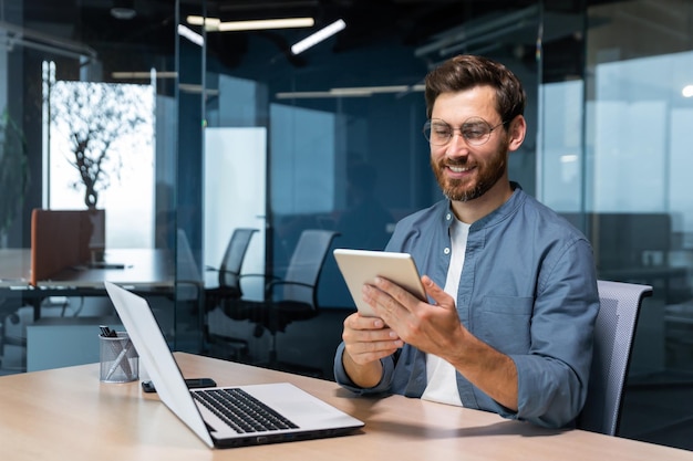 Un uomo d'affari di successo in una camicia usa un computer tablet un uomo si siede a una scrivania sorridendo e felice a