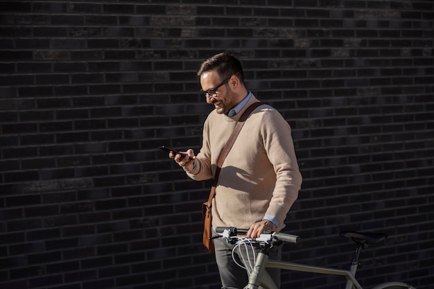 Un uomo d'affari felice che spinge una bicicletta e invia messaggi di testo mentre sorride al telefono