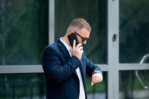 Un uomo d'affari fuori dall'edificio degli affari parla al telefono e guarda l'orologio