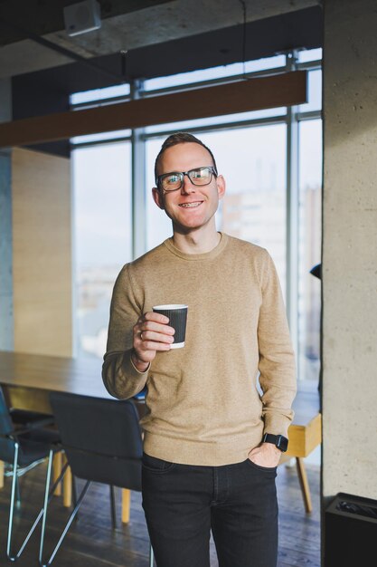 Un uomo d'affari in occhiali e abiti eleganti beve caffè in uno spazio di lavoro moderno Un libero professionista maschio con un laptop lavora da remoto