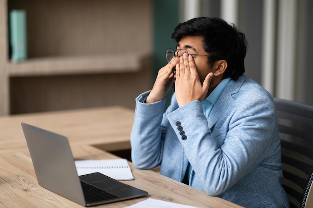 Un uomo d'affari indiano stressato al portatile strofinando gli occhi stanchi in ufficio.