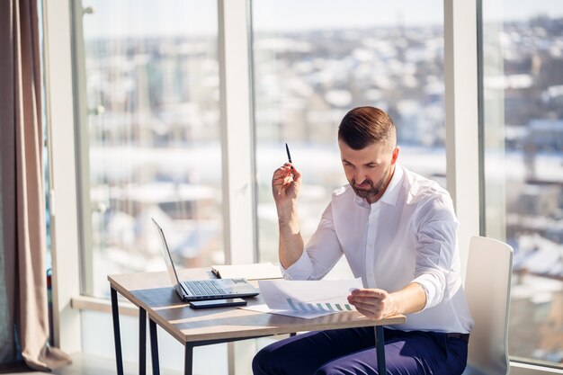 Un uomo d'affari maschio adulto di successo siede nel suo ufficio con grandi finestre e lavora a un laptop, lavorando a nuovi progetti.