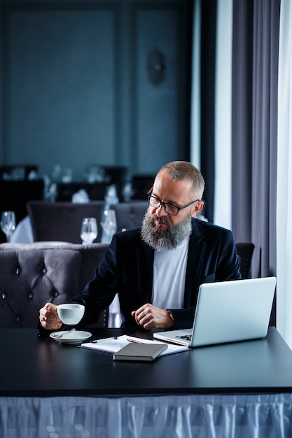 Un uomo d'affari maschio adulto sta lavorando a un nuovo progetto e sta guardando i grafici di crescita delle azioni. Si siede al tavolo vicino alla grande finestra. Guarda lo schermo del laptop e beve caffè.