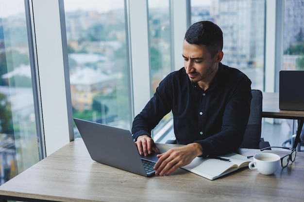 Un uomo d'affari sorridente con una camicia nera è seduto a una scrivania e utilizza un laptop Una persona lavora in ufficio su un computer analizza i dati Internet marketing educazione degli adulti elearning