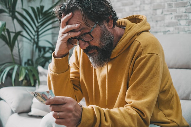 Un uomo da solo a casa con in mano una banconota in contanti che ha problemi economici tenendosi la fronte preoccupato e triste Perdere il lavoro Crisi in Europa Uomini preoccupati con soldi che pensano a una soluzione