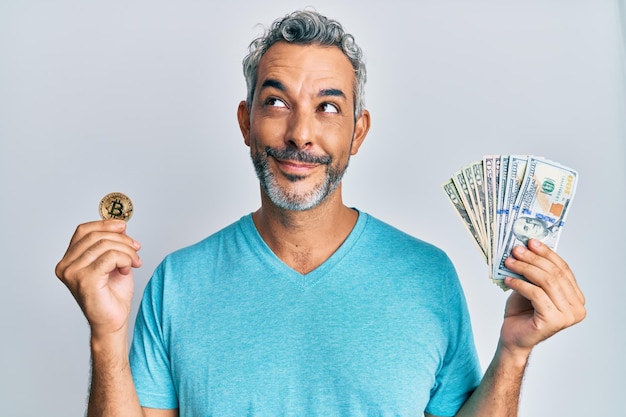 Un uomo dai capelli grigi di mezza età con in mano dollari e bitcoin sorride guardando di lato e distoglie lo sguardo pensando