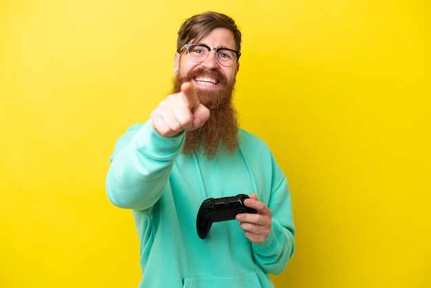 Un uomo dai capelli rossi con la barba che gioca con un controller per videogiochi isolato su sfondo giallo punta il dito contro di te con un'espressione sicura