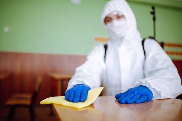 Un uomo del gruppo di disinfezione pulisce la scrivania a scuola con uno straccio giallo. Il lavoratore professionista sterilizza l'aula per prevenire la diffusione del covid-19. Assistenza sanitaria di alunni e studenti concetto.