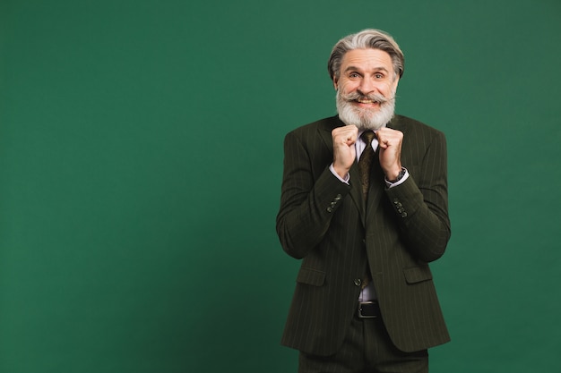 Un uomo di mezza età con la barba in giacca e cravatta tiene i pugni, fa male e si rallegra su una parete verde.