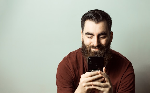 Un uomo di stile giovane hipster con barba e taglio di capelli moderno che sorride mentre guarda il suo schermo del telefono con spazio di copia