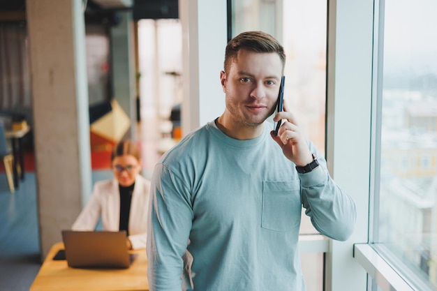 Un uomo di successo in abiti casual è in piedi vicino alla finestra nell'area di lavoro e parla al telefono Un giovane manager lavora a distanza