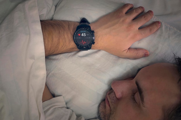 Un uomo dorme su un letto con un controllo della frequenza cardiaca nel sonno Gli orologi intelligenti misurano il polso sulla mano di una persona che dorme Fitness Activity Tracker con la frequenza cardiaca sulla mano dell'uomo sul letto