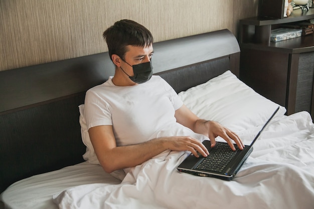 Un uomo è seduto a letto nella sua camera da letto e lavora a un laptop con una maschera medica sul viso