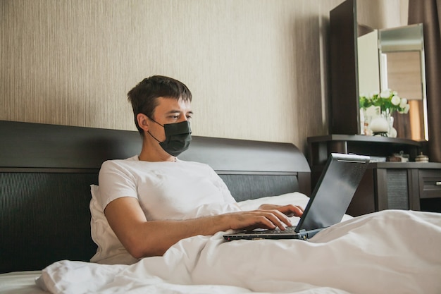 Un uomo è seduto a letto nella sua camera da letto e lavora a un laptop con una maschera medica sul viso