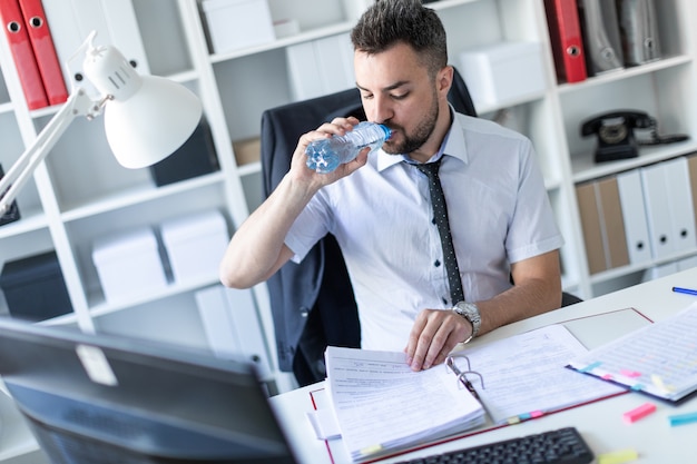 Un uomo è seduto a un tavolo in ufficio, lavora con documenti e beve acqua da una bottiglia.