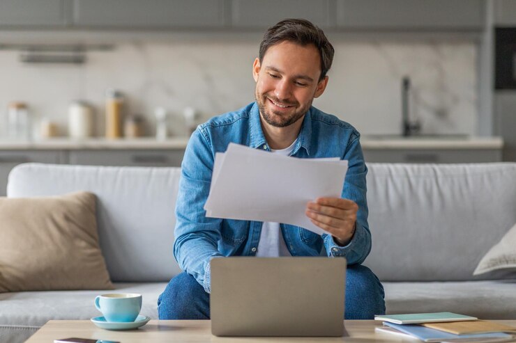 Foto un uomo è seduto su un divano sorridendo mentre rivede alcuni documenti mentre usa un computer portatile