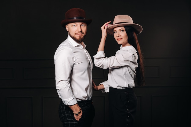 Un uomo e una donna in camicie bianche e cappelli su sfondo nero. Una coppia innamorata posa all'interno dello studio.