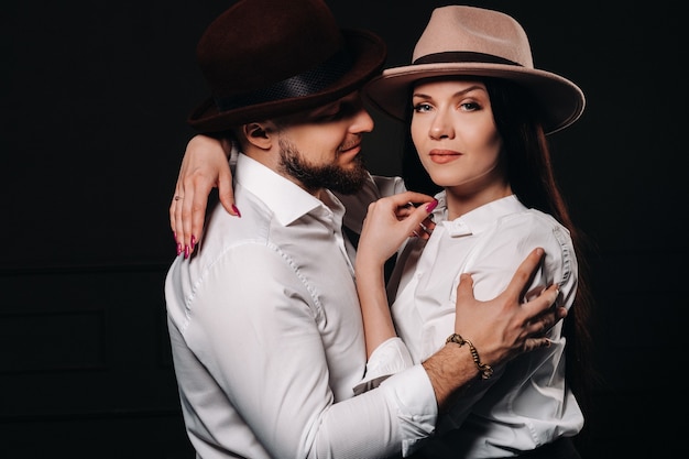 Un uomo e una donna in camicie bianche e cappelli su sfondo nero. Una coppia innamorata posa all'interno dello studio.