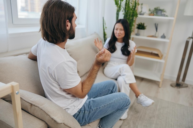 Un uomo e una donna si siedono sul divano di casa in magliette bianche e urlando comunicano scandalosamente non si capiscono Una lite in famiglia di due coniugi e aggressività emozioni tristi