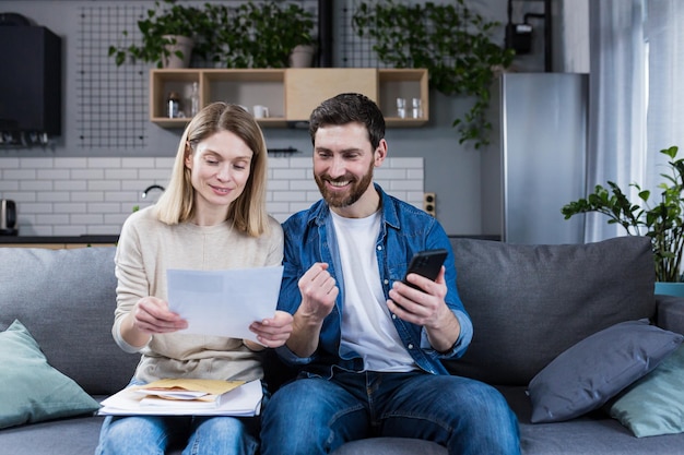 Un uomo e una donna sono felici a casa, effettuano pagamenti online utilizzando il telefono e l'applicazione bancaria seduti sul divano e tenendo le bollette