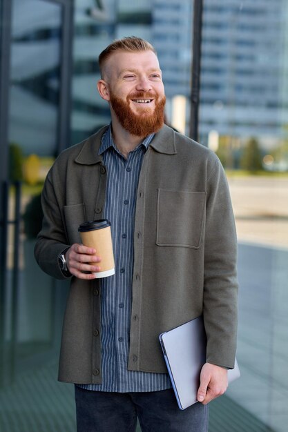 Un uomo europeo con la barba rossa vestito di camicia e giacca cammina per la città e beve caffè durante la pausa pranzo