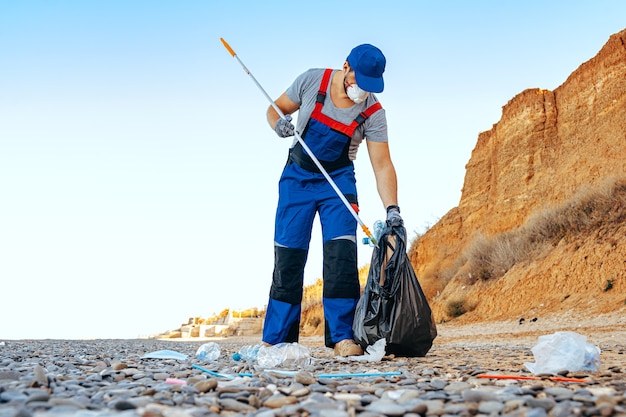 Un uomo fa volontariato in uniforn raccogliendo spazzatura sulla spiaggia con un estensore di portata