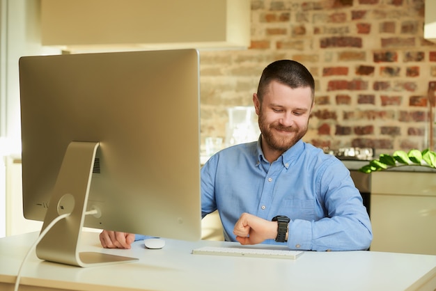 Un uomo felice con la barba che guarda l'orologio da polso durante un video briefing online con i suoi colleghi a casa