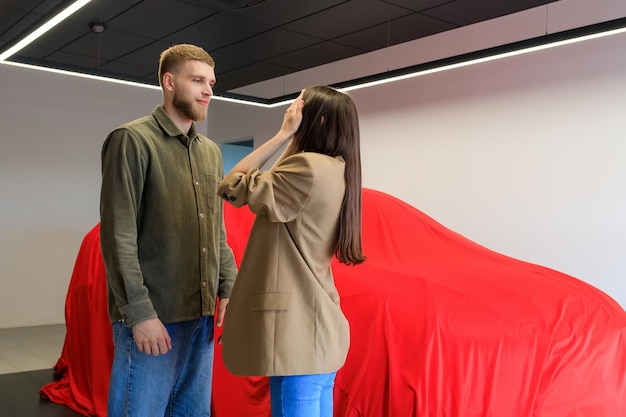 Un uomo ha fatto un regalo a una donna comprando un'auto in una concessionaria coperta da un telo rosso Una giovane donna si copre gli occhi con le mani