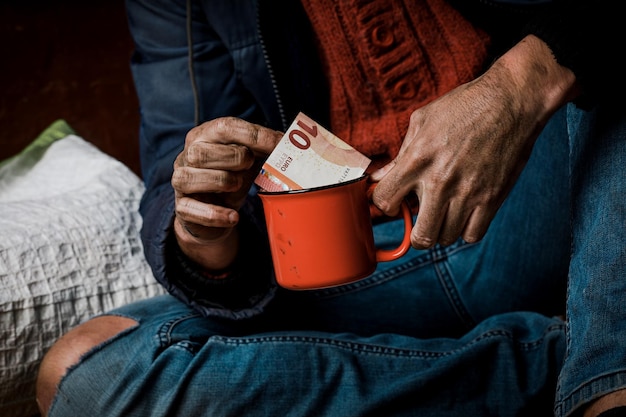 Un uomo implora. Ha in mano una tazza di carta bianca. Il concetto di senzatetto e povertà.