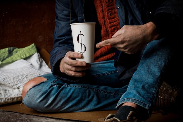 Un uomo implora. Ha in mano una tazza di carta bianca. Il concetto di senzatetto e povertà.