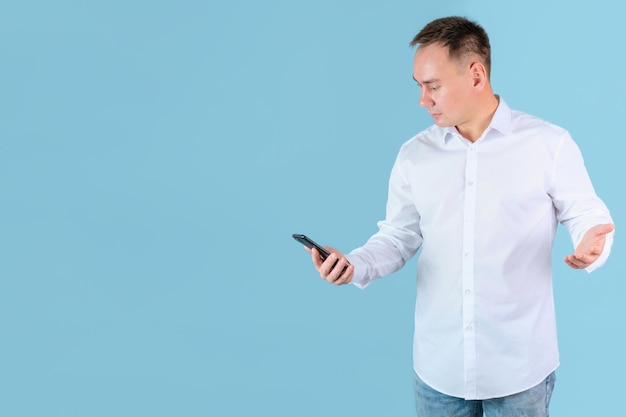 Un uomo in camicia bianca guarda lo schermo del telefono e allarga irritato le mani. Sfondo blu, copyspace