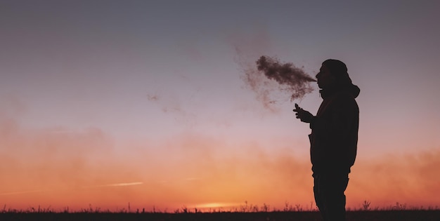 Un uomo in giacca fuma una sigaretta elettronica all'aperto Vaping su uno sfondo di tramonto