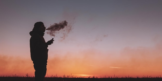 Un uomo in giacca fuma una sigaretta elettronica all'aperto Vaping su uno sfondo di tramonto
