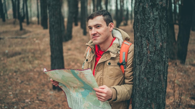 Un uomo in viaggio con mappa nei boschi Un ritratto del bell'uomo con uno zaino in piedi vicino a un albero in una stagione fredda